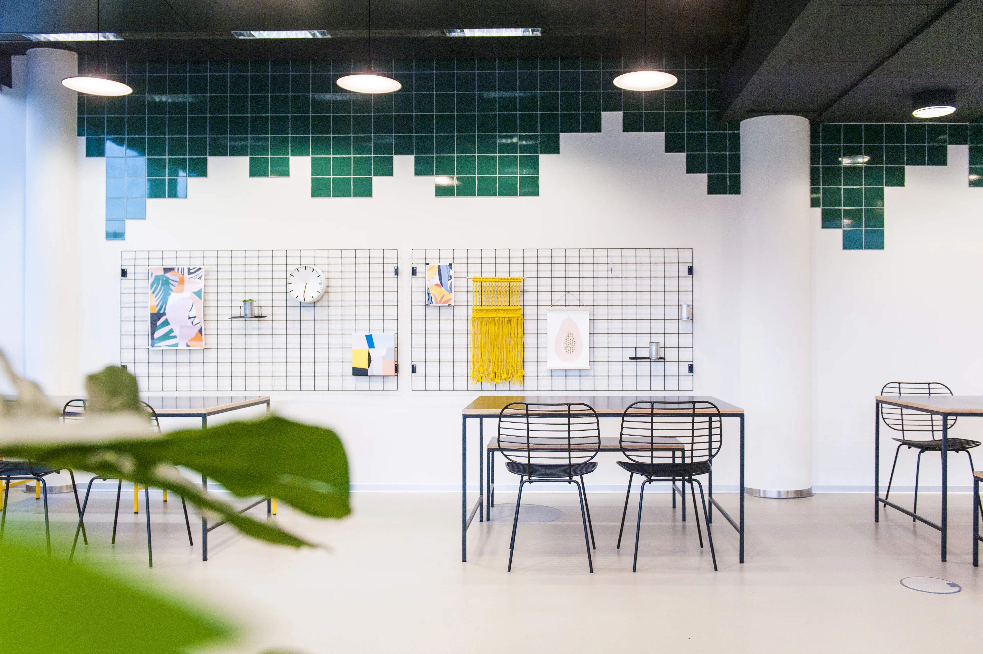 Seating area at KANTINE KOMTUR with black wire chairs at a wooden-edged table, wire grid wall panels with posters and a yellow macramé hanging, framed by green pixel tiles.