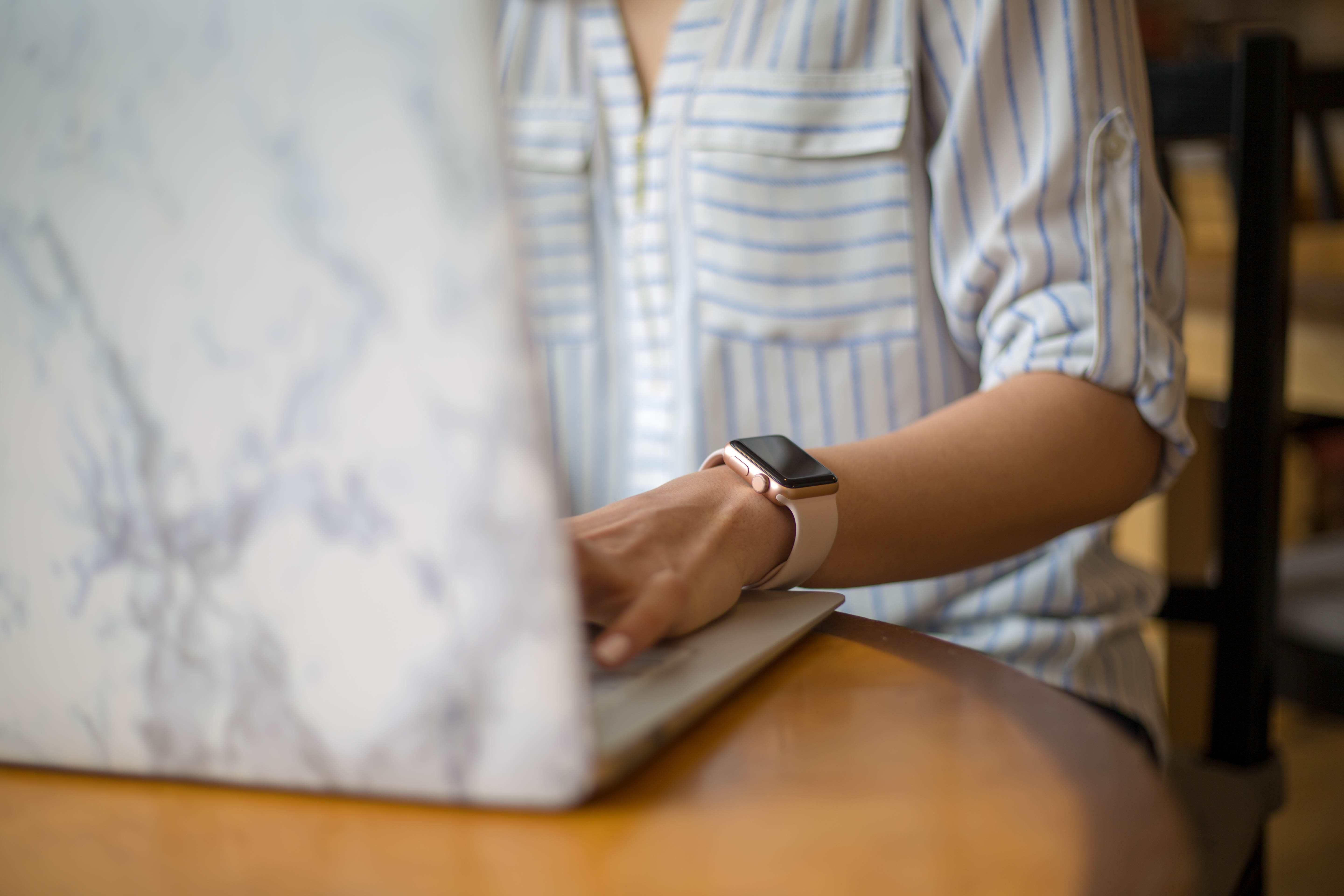 A person wearing a striped shirt and a smartwatch, working on a laptop with a marble-patterned cover, seated at a wooden table.