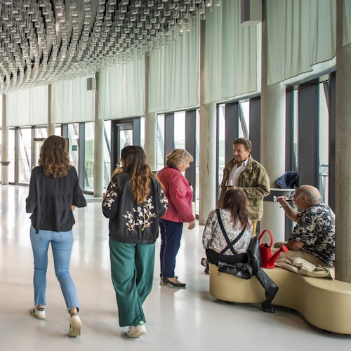 ผู้คนสนทนาและเดินในโถงทางเดินที่สว่างสดใสและทันสมัย มีหน้าต่างสูงจากพื้นจรดเพดานและการตกแต่งเพดานที่สวยงาม