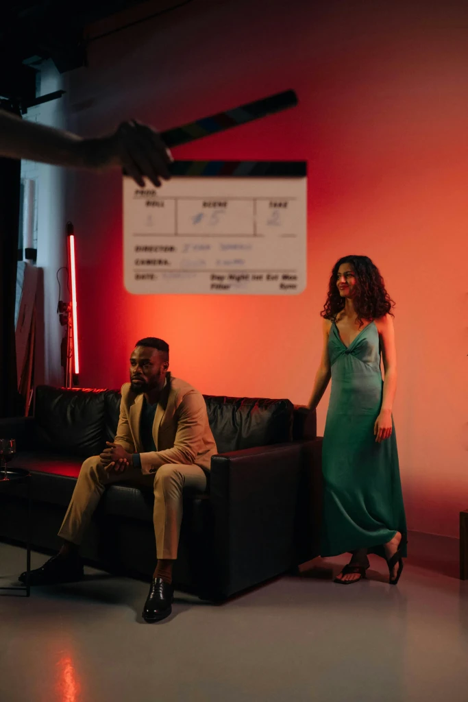 Man in a tan suit on a sofa and a woman in a green dress on a film set with red light. Blurry clapperboard in foreground.
