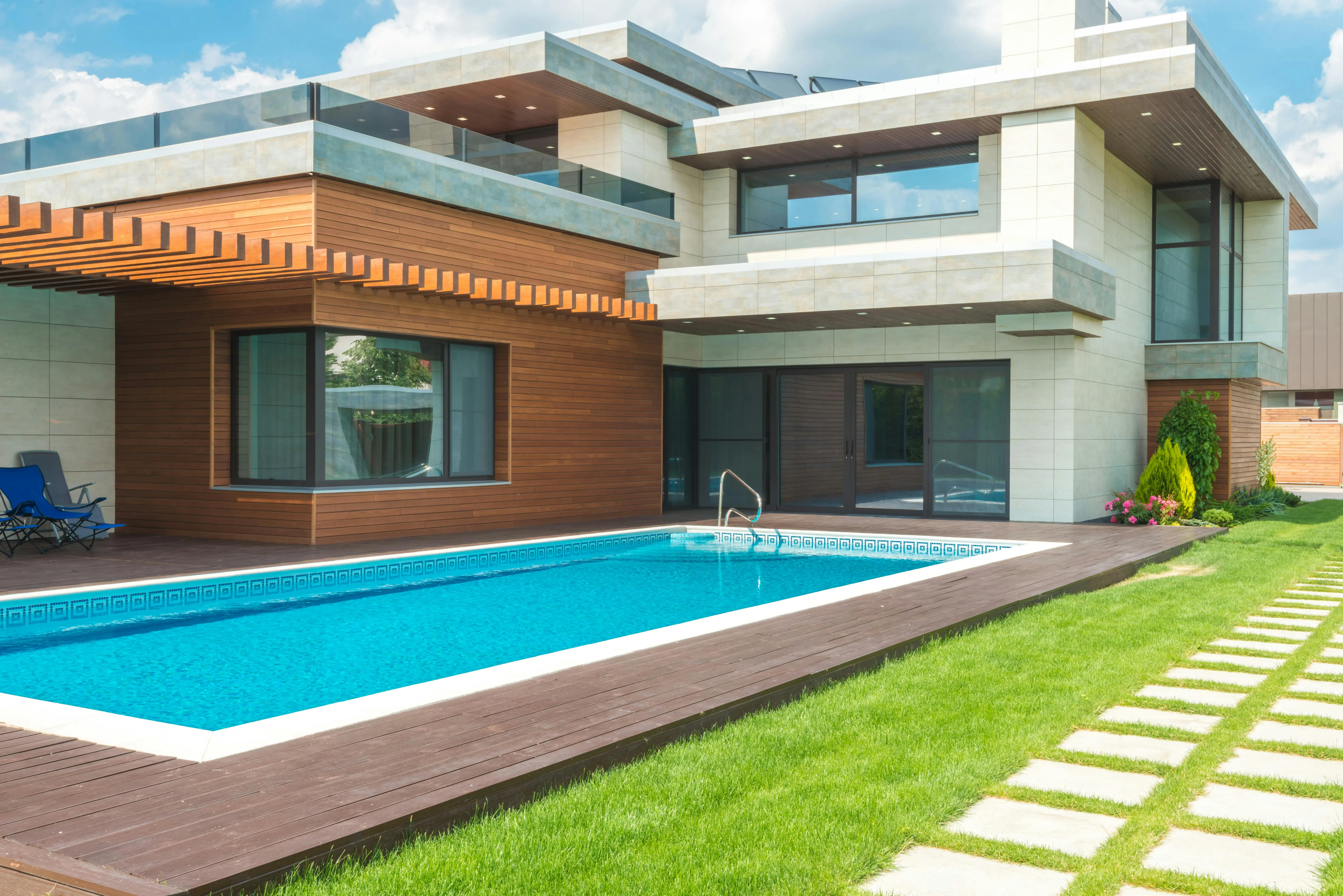 Modern house with a flat roof, wood and concrete facade, and large windows. A clear blue swimming pool with wooden deck is surrounded by green lawn.