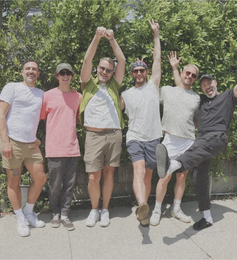 Group of six men laughing and posing playfully in front of green bushes at an AwareHaus gathering.