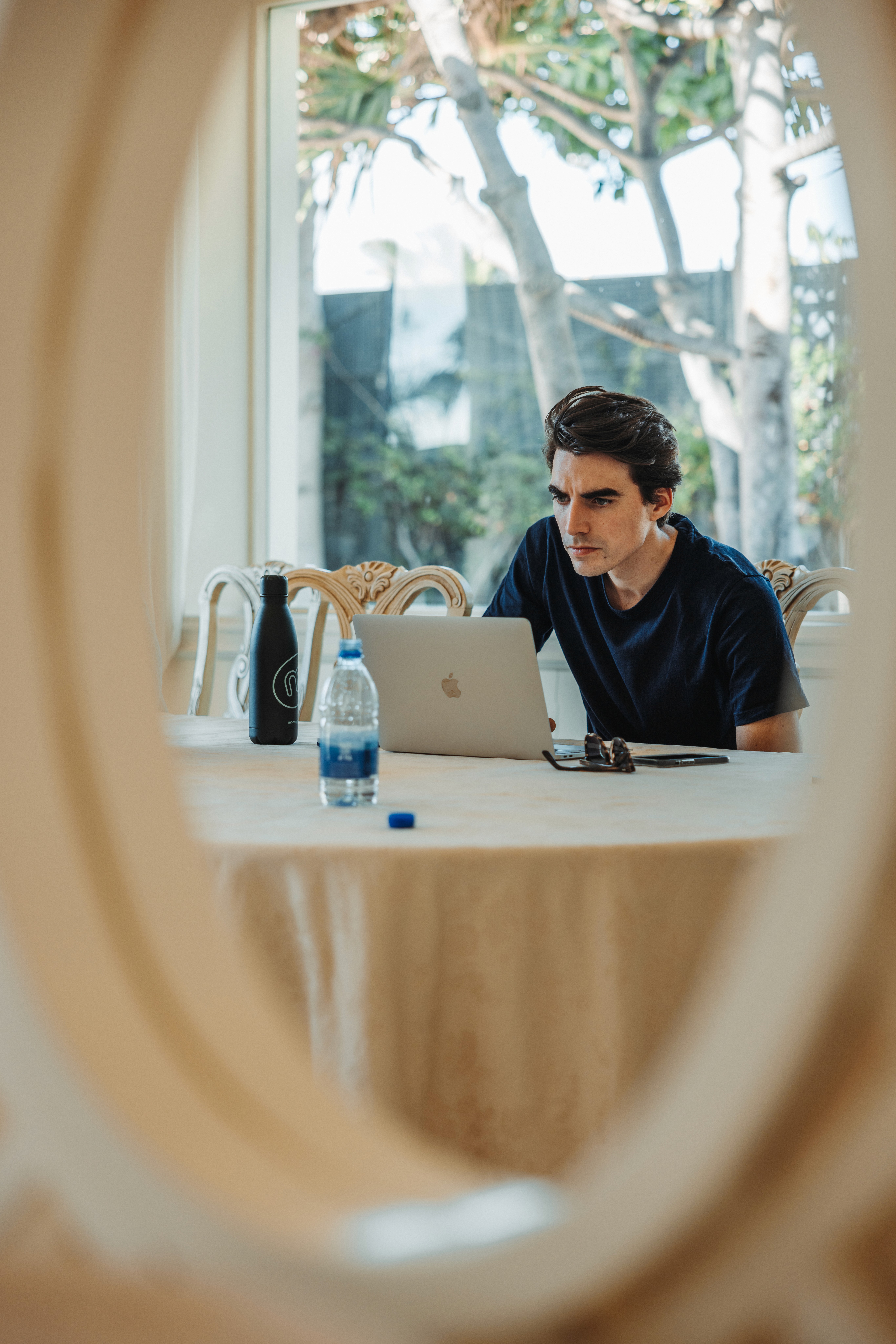 Person working on a laptop at a table with two bottles, sunglasses, and a smartphone nearby