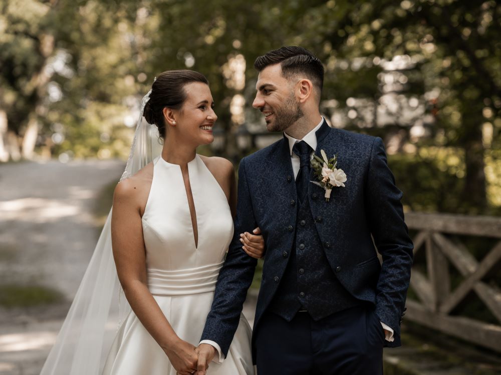 Kundenfeedback Hochzeitspaar für Maria Wiethaler freie Rednerin und Sängerin. Strahlendes Brautpaar an ihrem Hochzeitstag: Die Braut in einem eleganten weißen Hochzeitskleid mit tiefem Ausschnitt und langem Schleier, der Bräutigam in einem stilvollen dunkelblauen Anzug mit floraler Ansteckblume. Sie lächeln sich verliebt an, während sie Hand in Hand durch eine grüne, romantische Umgebung spazieren. Hochzeitsfotografie mit natürlichem Licht und authentischen Emotionen.