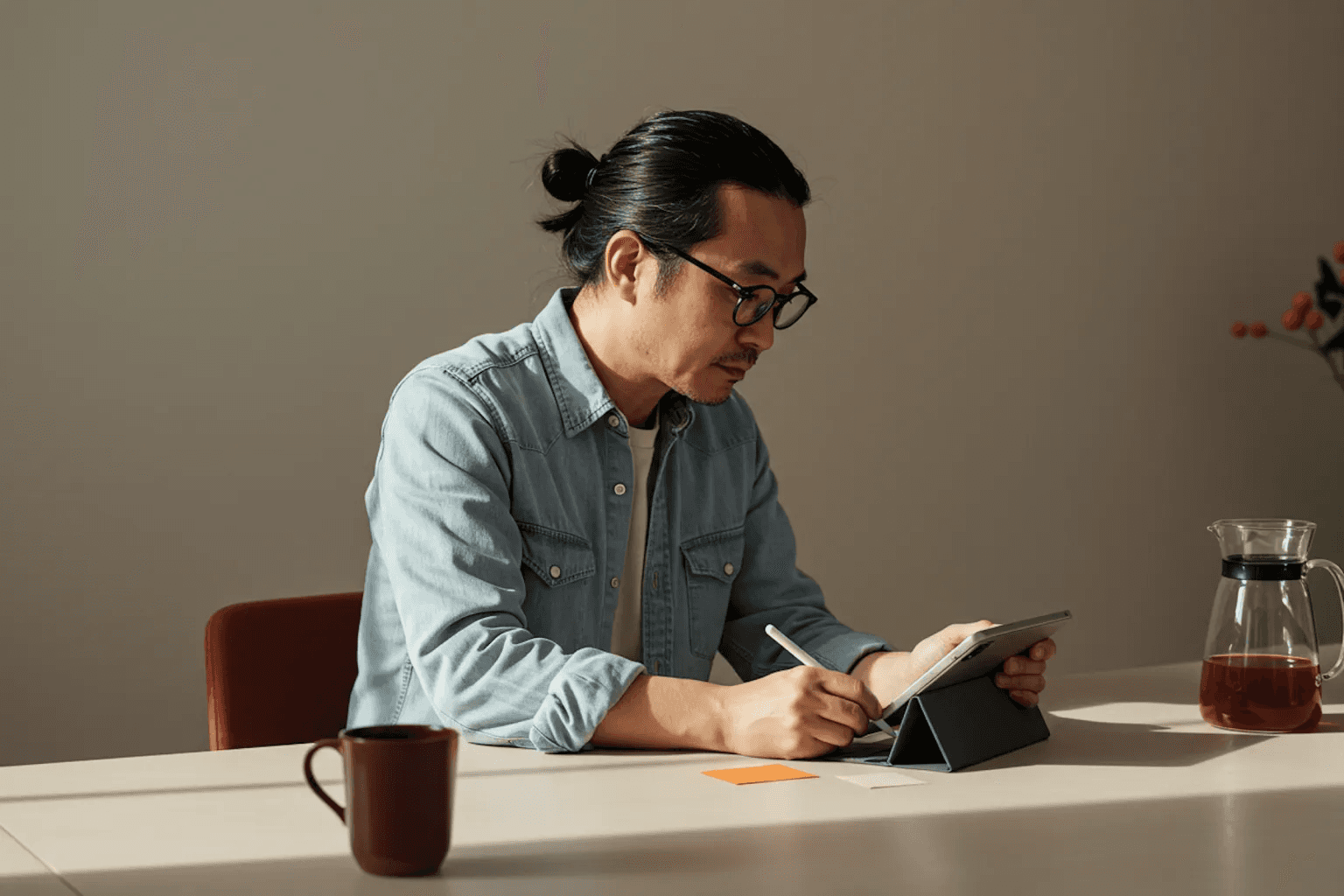 Man with glasses and tied-back hair writing at a desk while looking at a tablet.