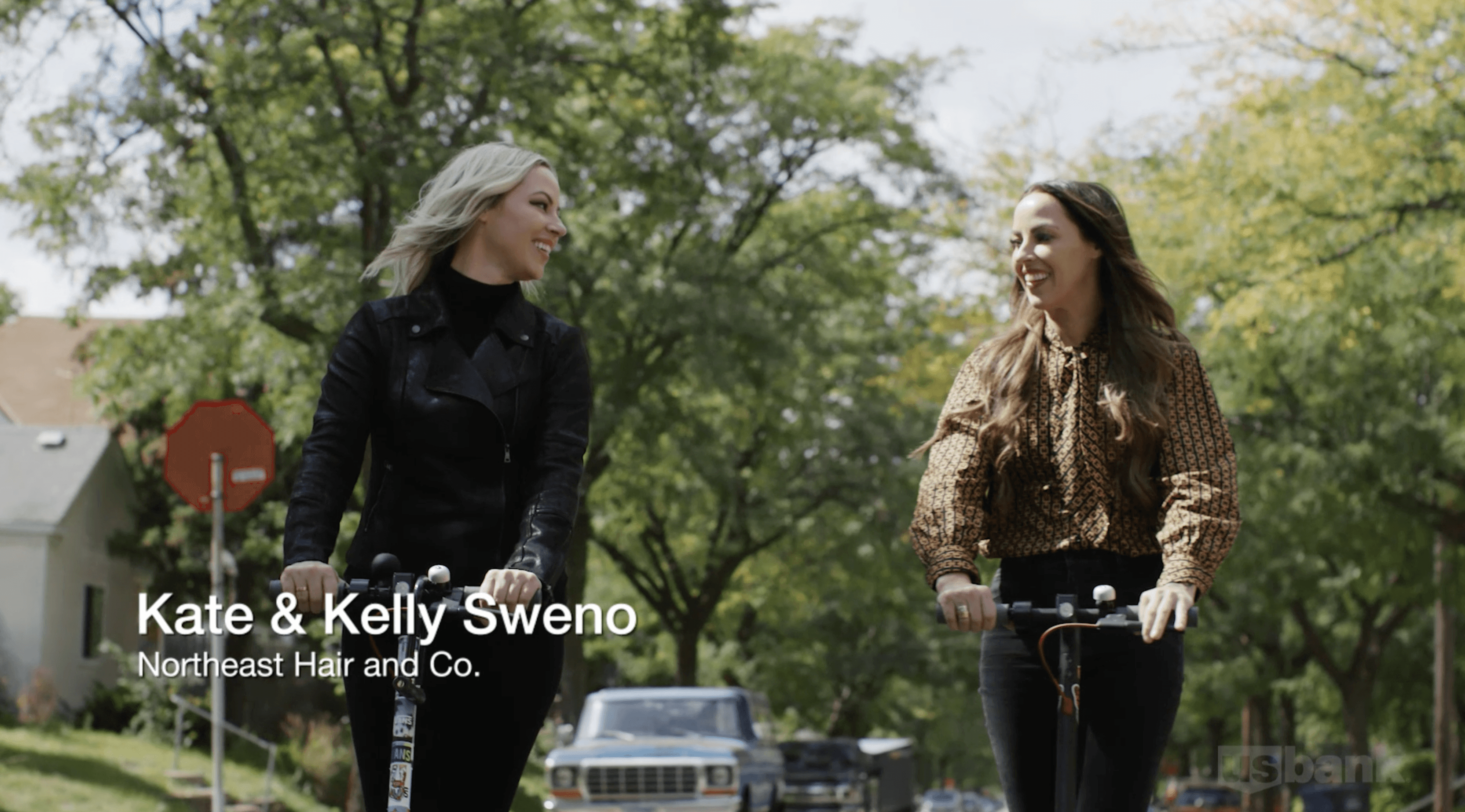 Two sisters smile at each other as they scooter to work, one is blonde and looks edgy, the others is a brunette and looks more relaxed.
