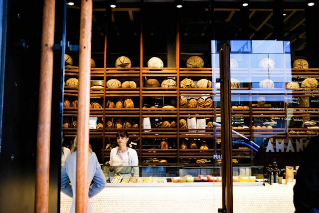 This image shows a woman, possibly a bakery owner or staff member, standing behind a counter filled with pastries and bread in a bakery with shelves of neatly arranged loaves behind her. This image serves as the hero image for the blog on the Rarecide website, discussing 'The Impact of Social Media on Customer Acquisition and Retention.' Rarecide, a top-rated social media management and marketing agency, specializes in social media marketing, marketing strategy development, and customer engagement. Known as the best marketing agency in Toronto, Rarecide excels in innovative marketing strategies for business growth.