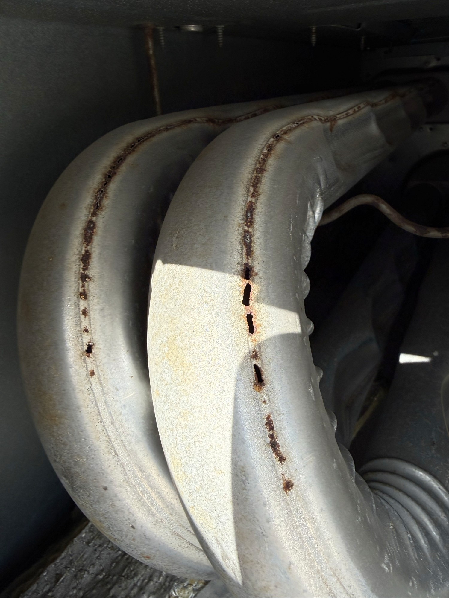 Close-up of a cracked heat exchanger inside a rooftop furnace, with rust-through holes visible along the seam of the metal tube.