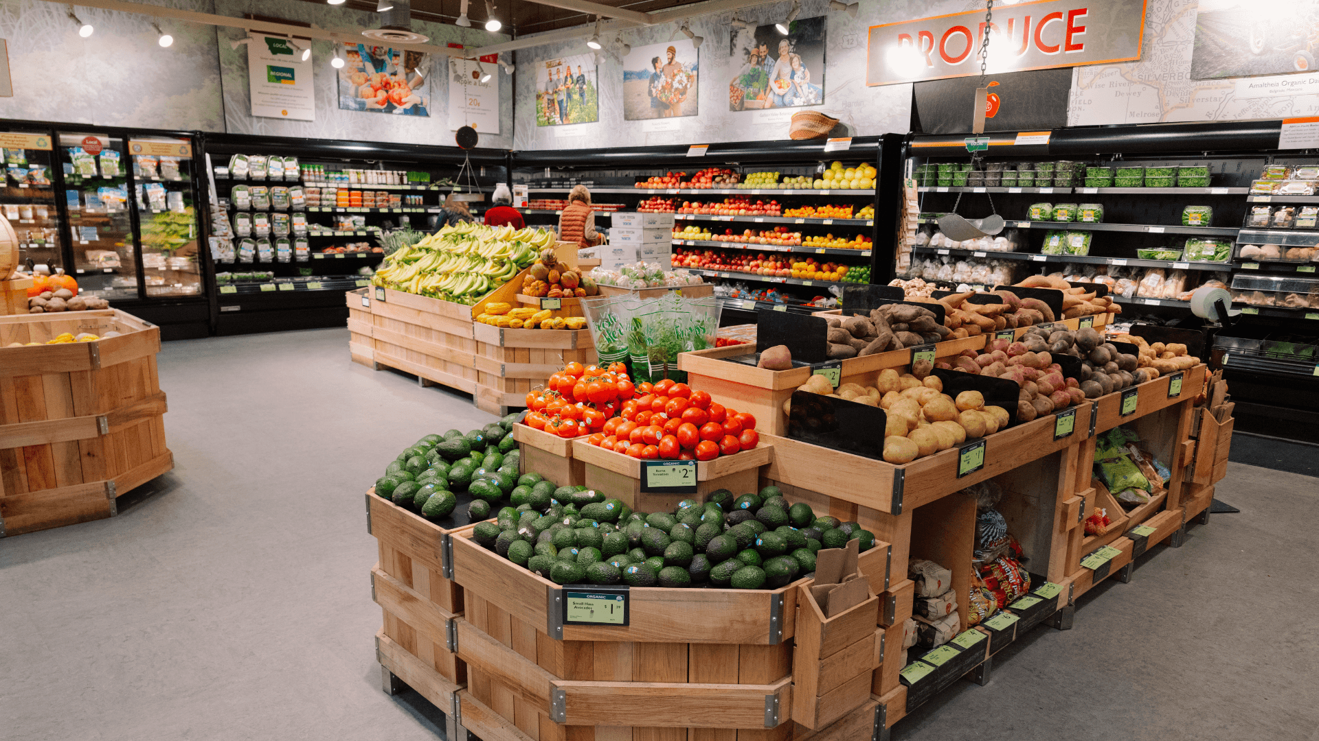 A store employee in an apron holds up two items in front of a colorful produce display.