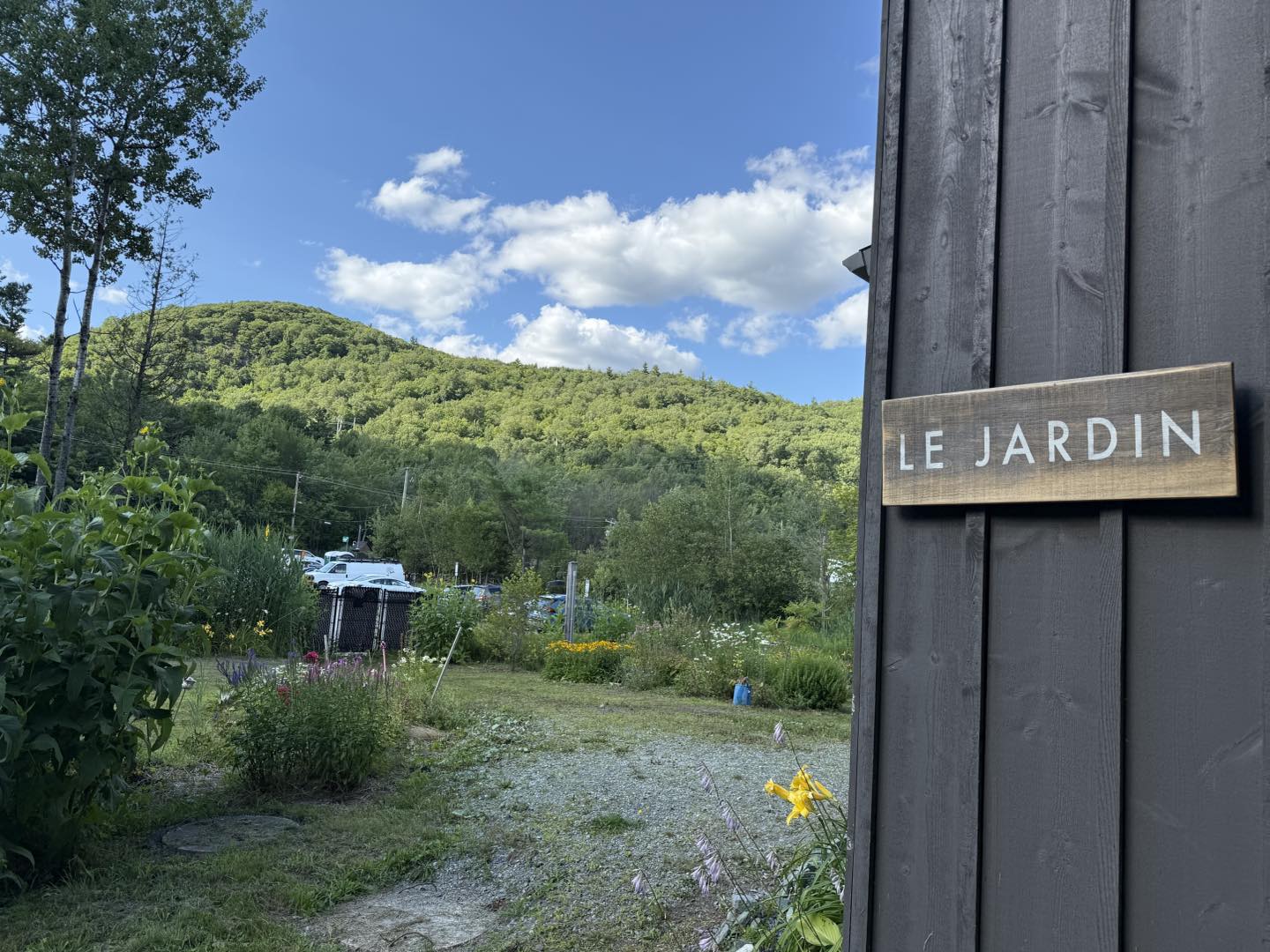 Jardin du gîte R112 Nord à Austin, avec vue sur les montagnes verdoyantes sous un ciel bleu parsemé de nuages, panneau en bois « Le Jardin » au premier plan.