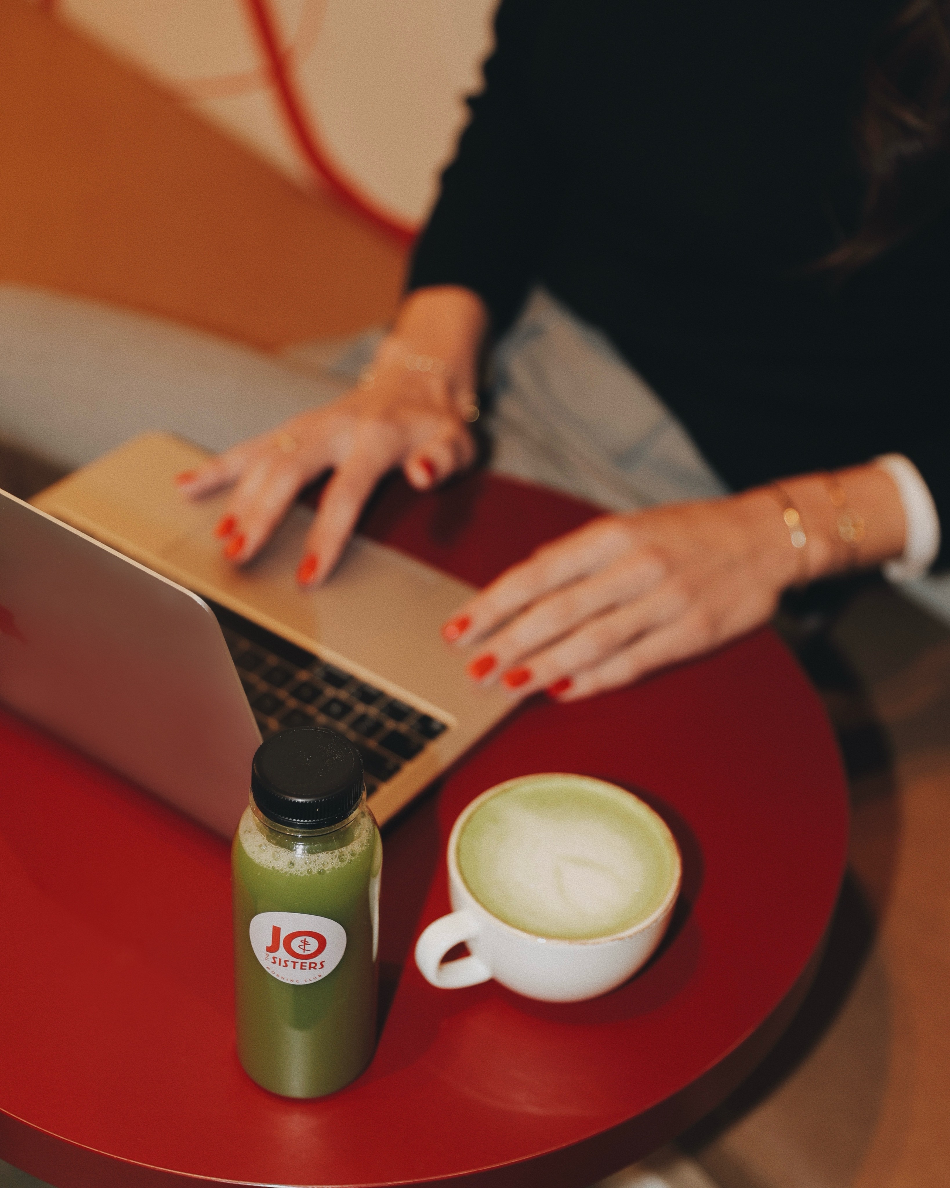 Person working on a laptop with a matcha latte and green juice on a red table at Jo & The Sisters Morning Club.