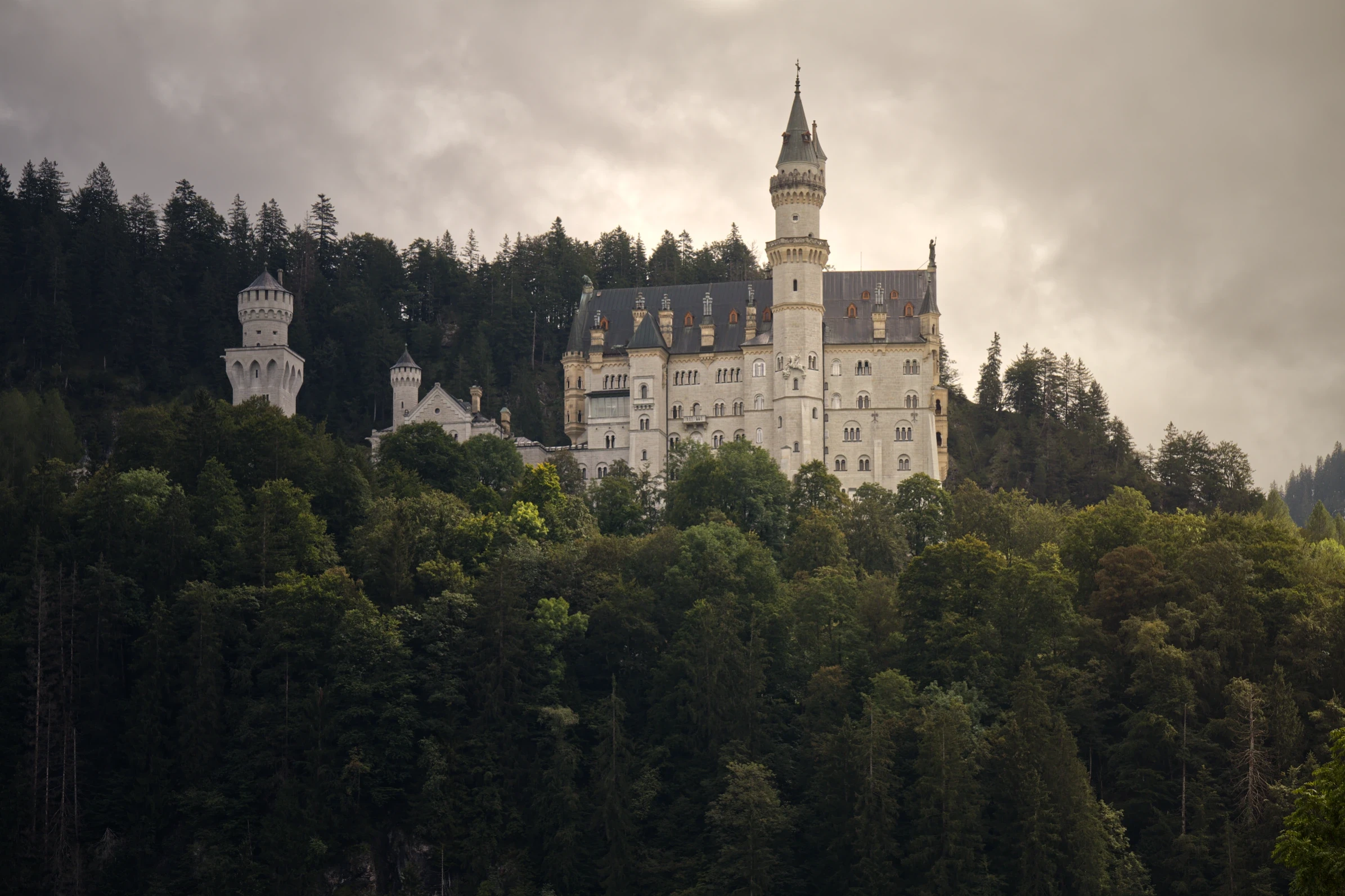 Historische Architektur von Schloss Neuschwanstein vor bewaldeten Berghängen und Wolkenformationen.