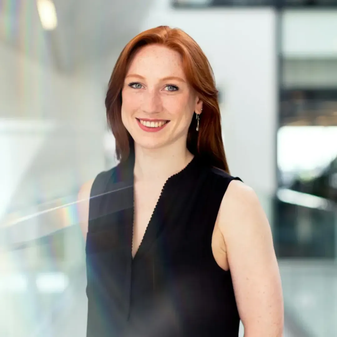 A woman with red hair and a black sleeveless top smiling confidently in a bright, modern indoor setting.