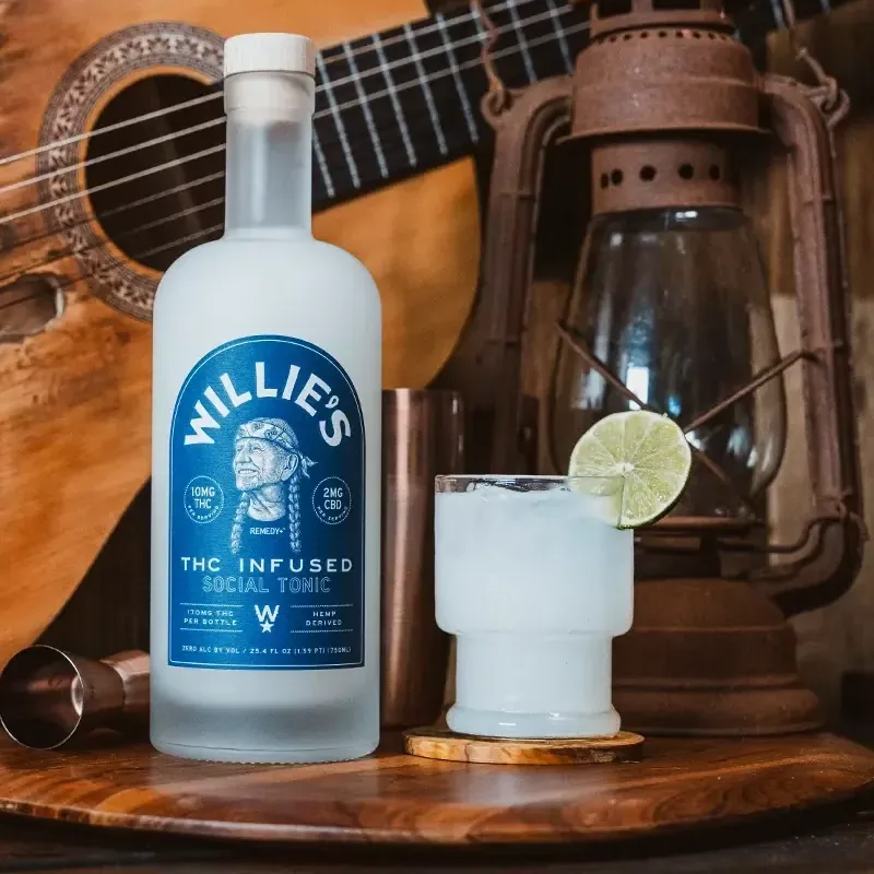 Close-up of a Willie’s Remedy+ social tonic bottle beside a cocktail garnished with lime and a rusty lantern