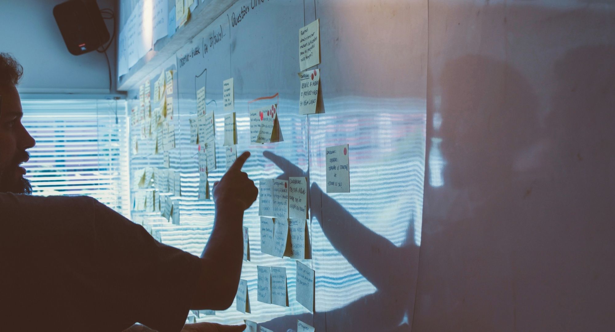 quadro branco com vários papéis "post-it" colados, iluminação baixa em tons de azul e amarelo um pouco refletida no quadro, um homem aponta para um dos papéis
