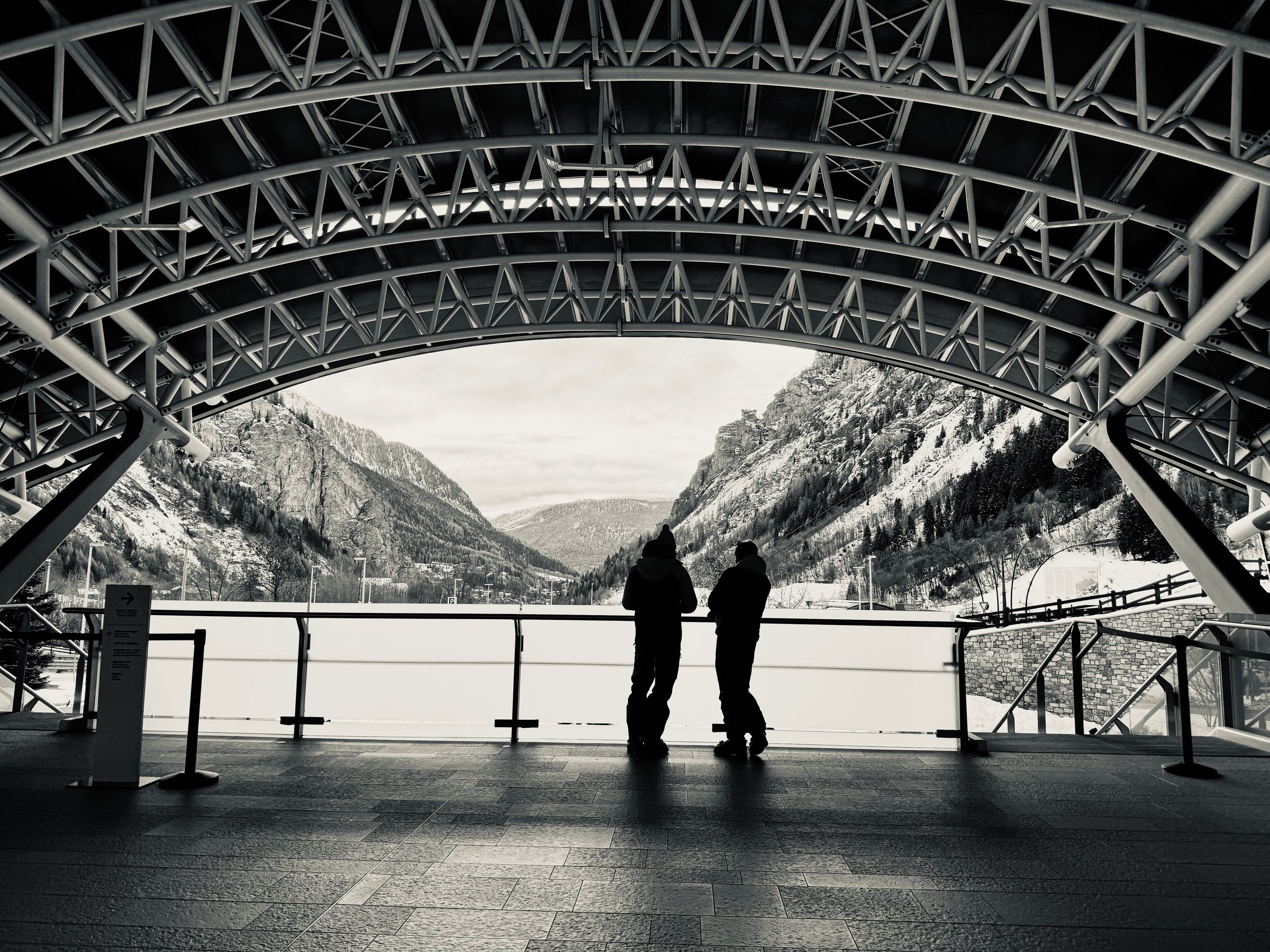 Two silhouetted figures under the arched steel canopy of the Skyway Monte Bianco station, Courmayeur
