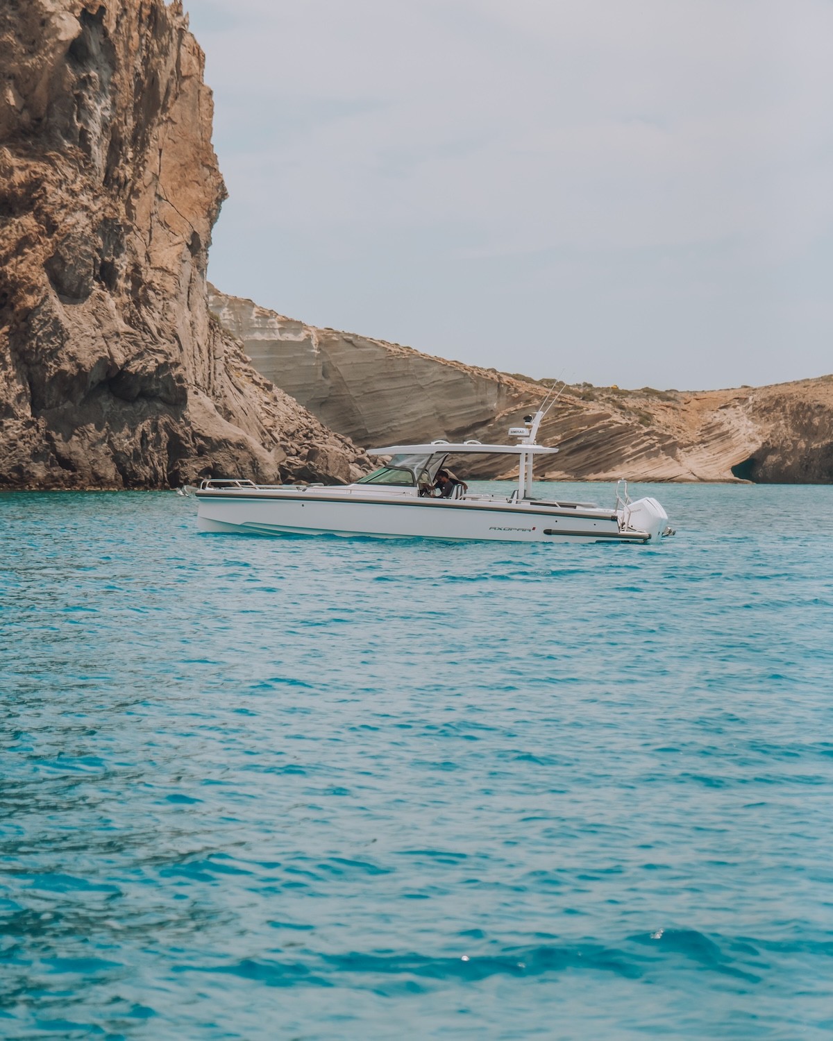 White Axopar 37 motor yacht cruising in crystal-clear turquoise waters alongside dramatic layered rock cliffs in the Cyclades.