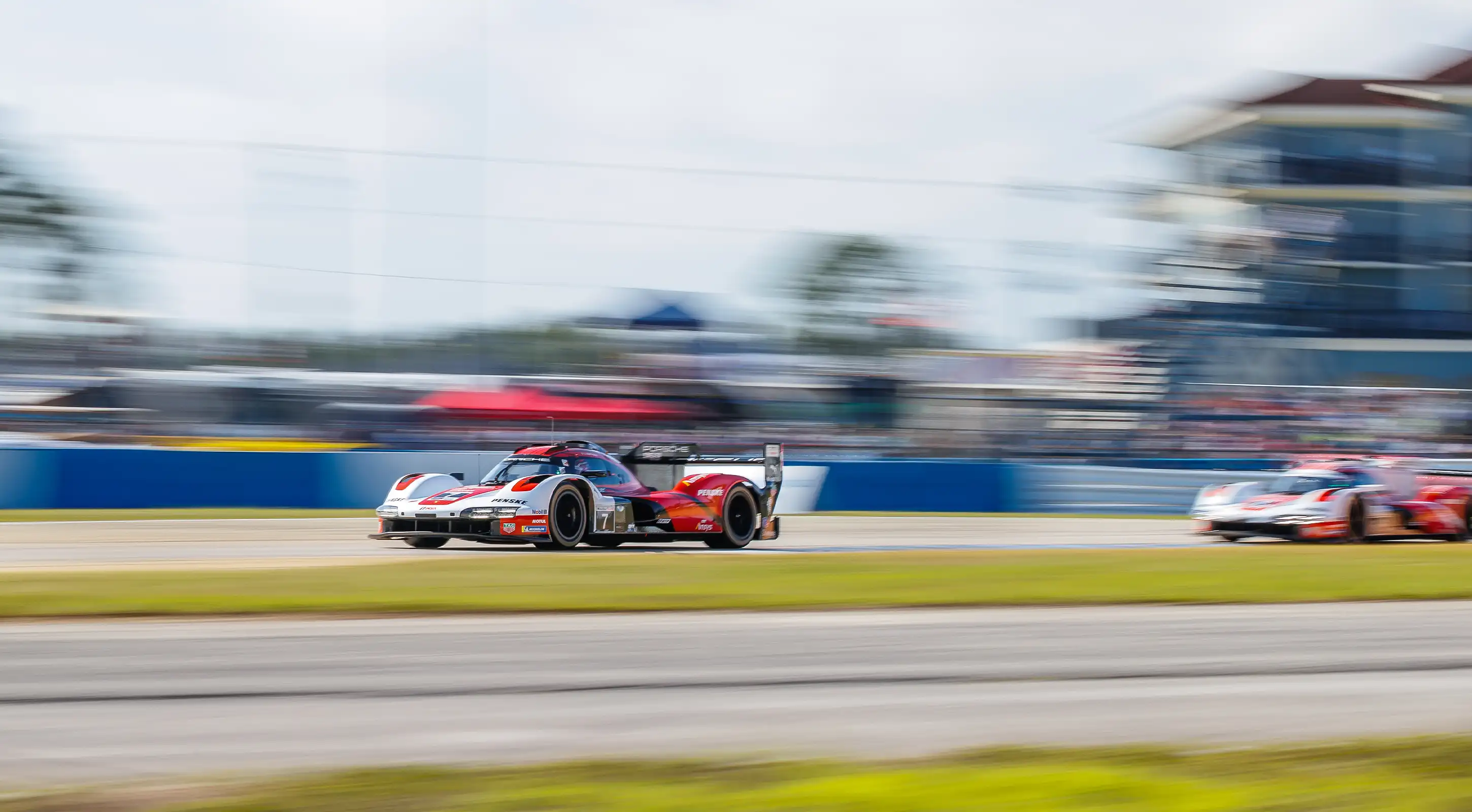 Two red, black, and white race cars compete on the Sebring track, one chasing the other, with a motion-blurred background emphasizing the intense speed.