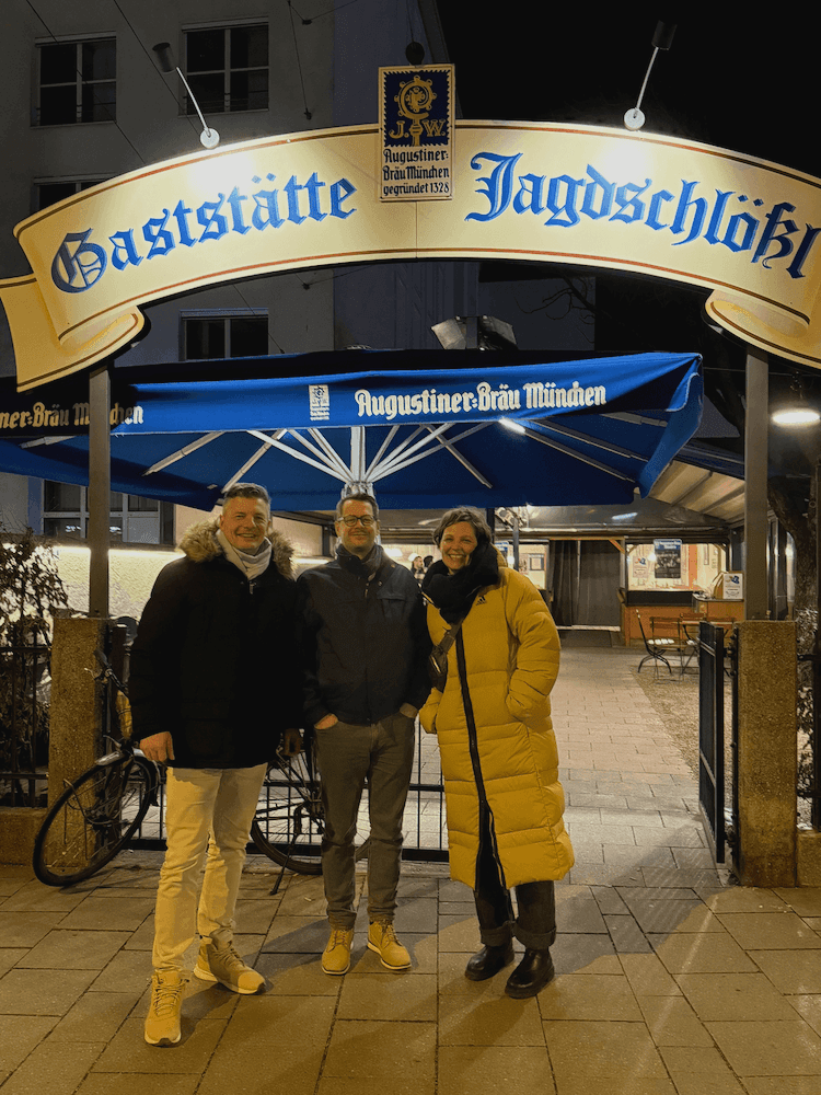 Drei Personen stehen vor dem beleuchteten Eingangsschild der Gaststätte Jagdschlößl mit blauem Augustiner-Bräu-München-Sonnenschirm im Hintergrund