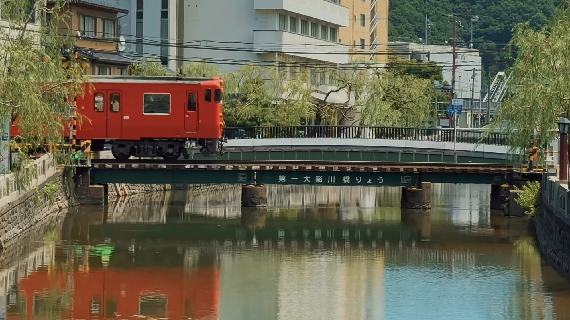 A red railcar crossing the girder bridge over the Otani river beside Kinosaki in summer.