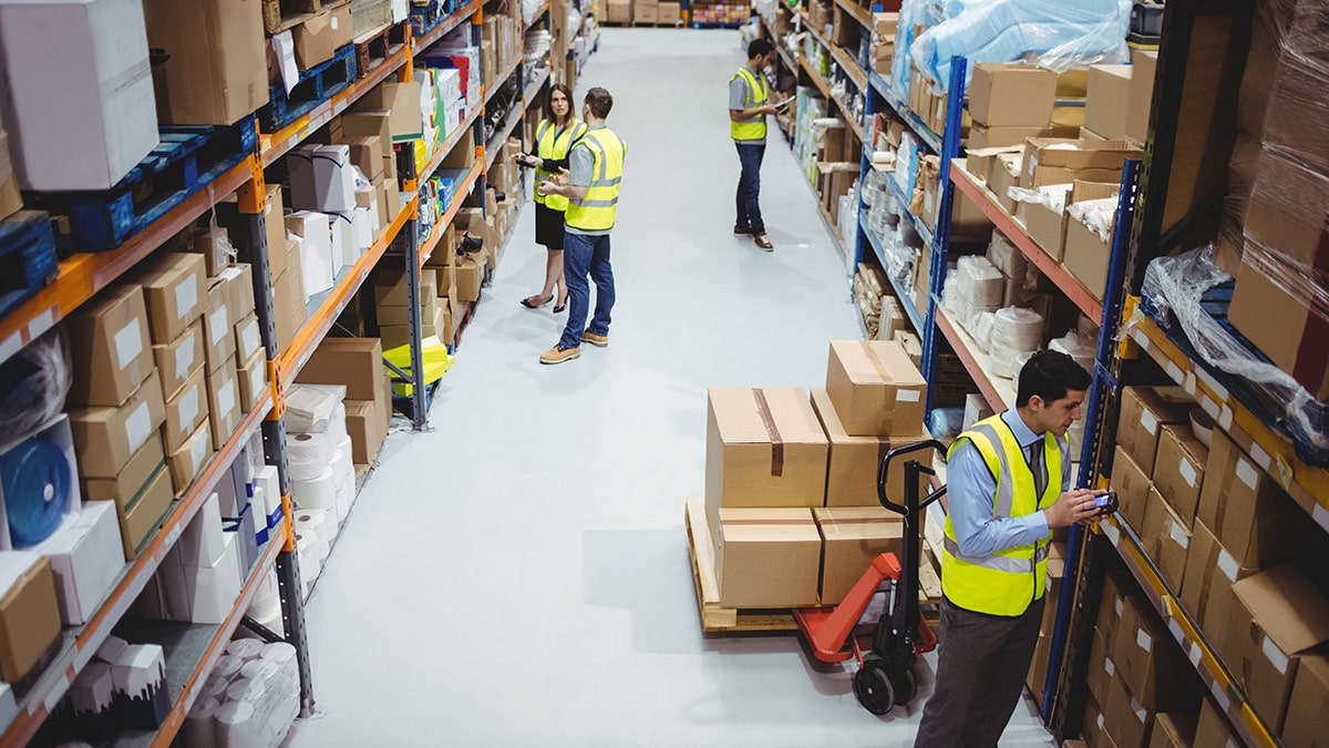 Workers in safety vests organize and handle boxes in a busy warehouse setting with shelves lined with goods.