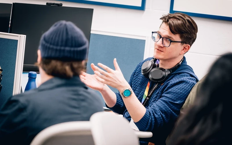 Person wearing a blue hoodie and headphones around the neck gestures with both hands while seated in an office setting, with computer monitors and partition panels in the background.
