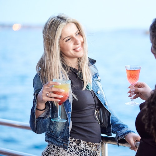 Two women enjoy colorful drinks on a boat, with one smiling and holding a cocktail. The ocean is in the background.