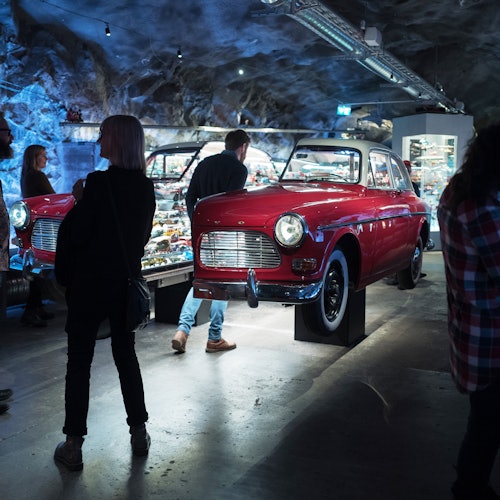Personer som observerar en röd klassisk bil som visas inomhus med rockiga dekorationer och olika mindre utställningar i bakgrunden.