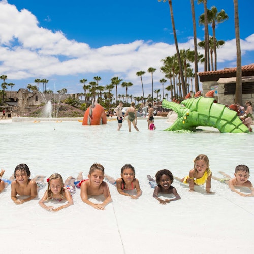 Children lying on their stomachs in a shallow pool with playful water features and palm trees in the background.