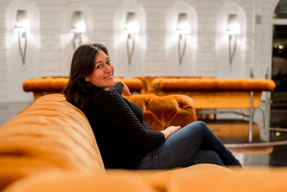 A woman in a black sweater and jeans smiles while sitting on an orange velvet sofa against a white arched wall.
