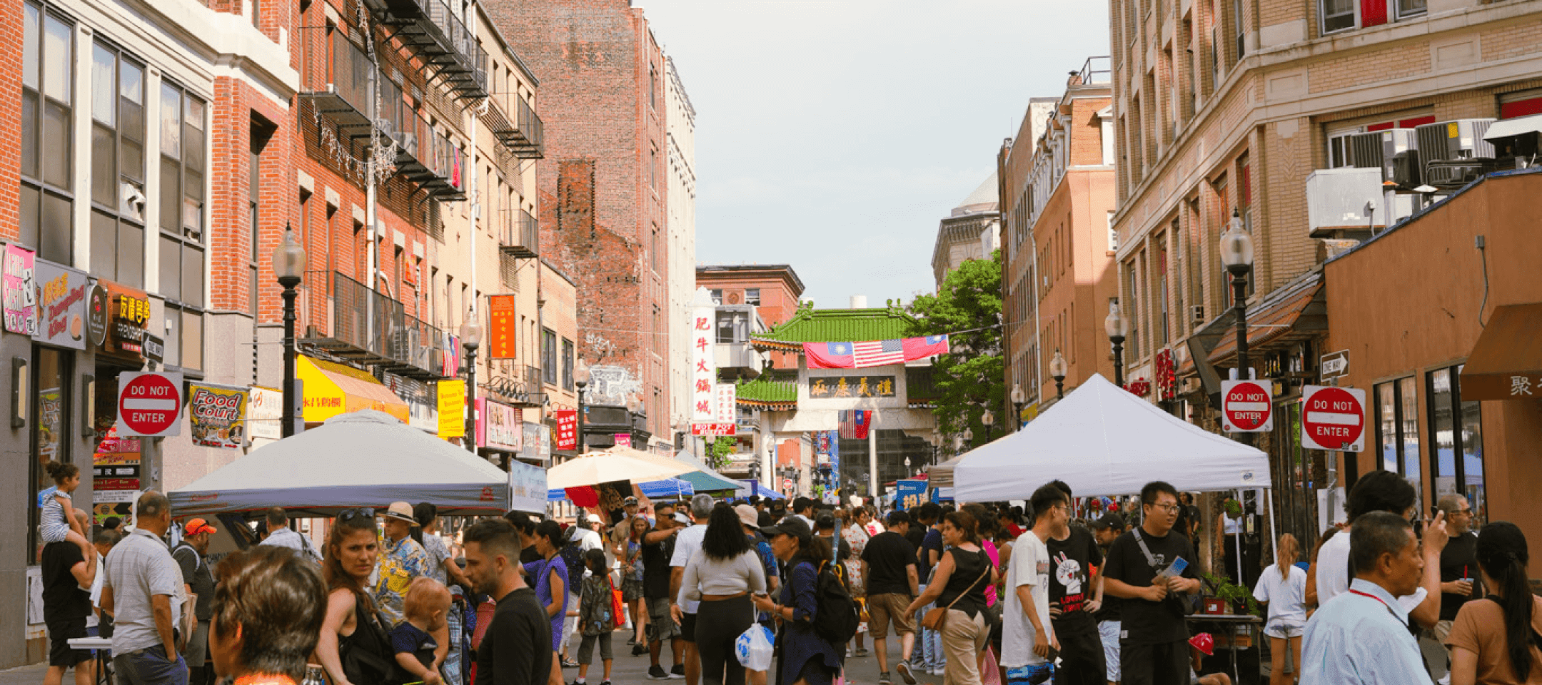 Crowds fill a street festival in Boston Chinatown, lined with vendor tents and colorful signs, with the Chinatown gate and flags visible in the background.