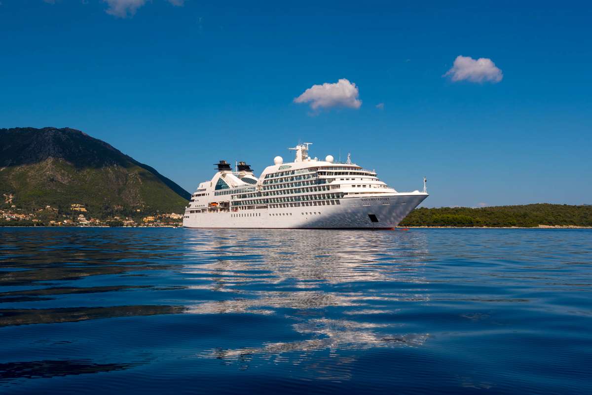 A large white cruise ship sails on blue water near a green coastline and mountains under a clear sky with clouds.