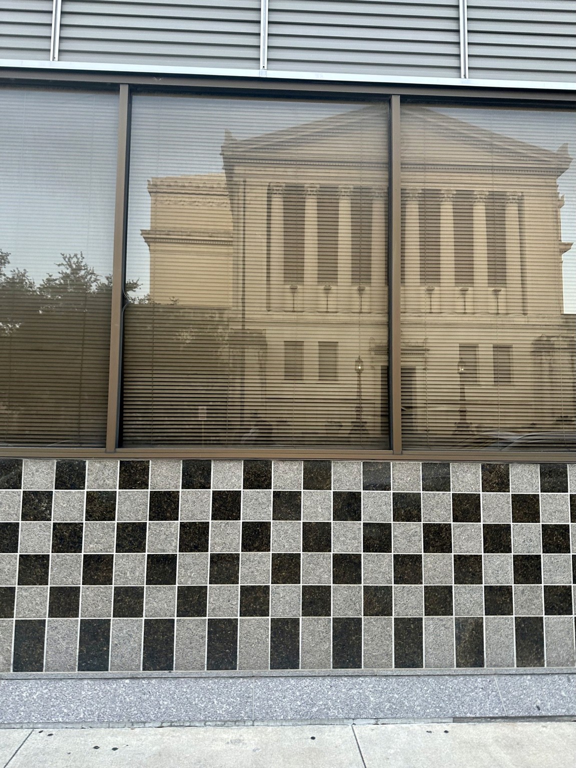A classical columned building reflected in glass above a black and white checkerboard wall.