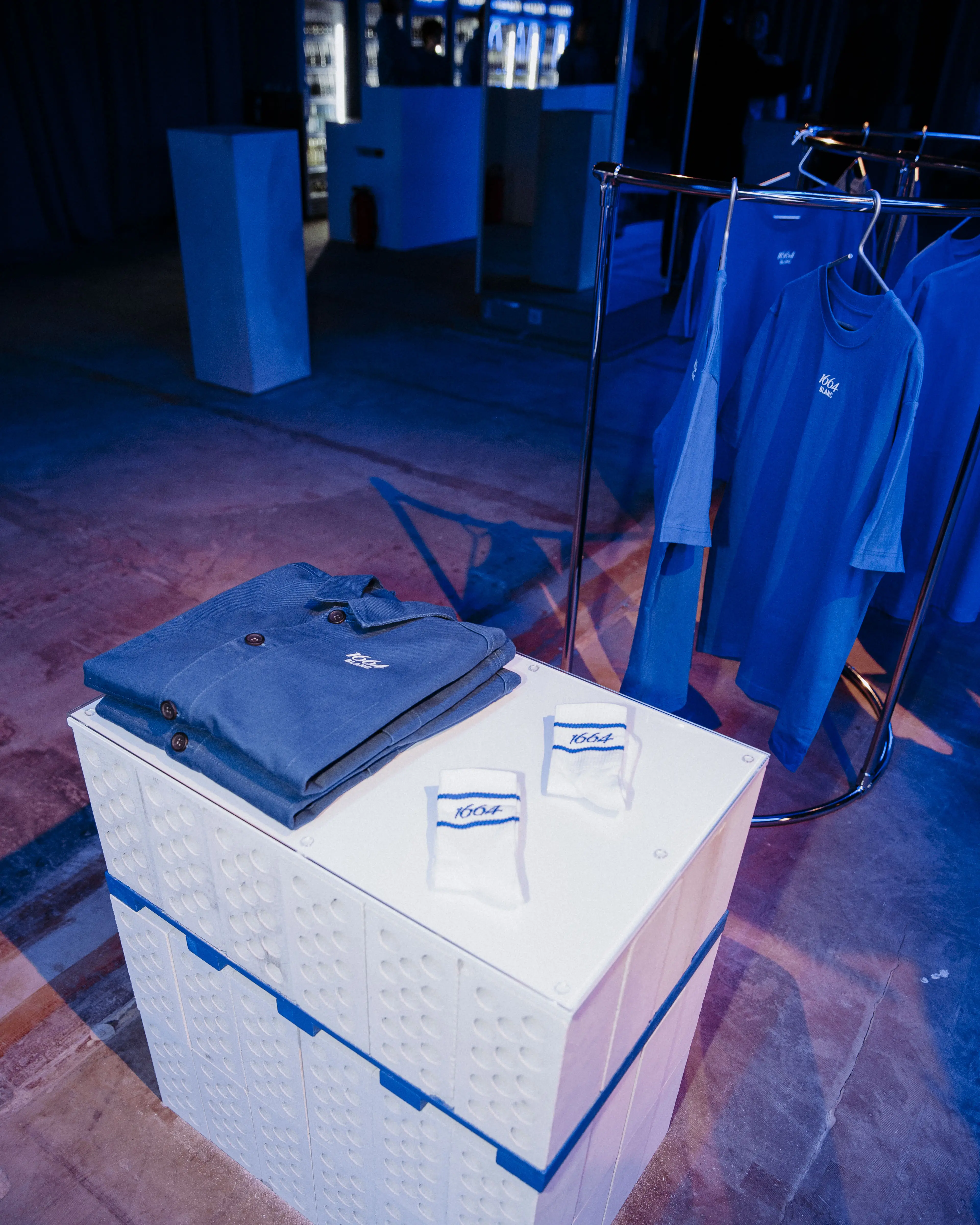 Folded blue polo shirt on white crate plinth next to a rack of 1664 Blanc shirts.