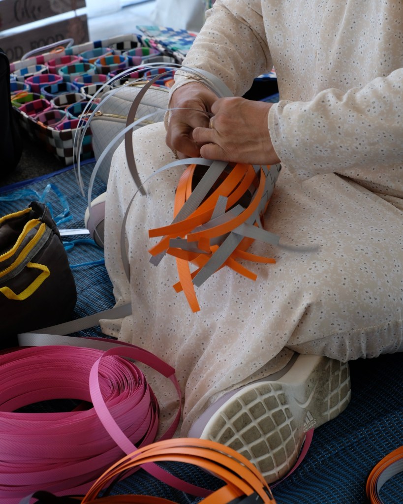 A close-up of hands skillfully weaving colorful strips of material, with vibrant rolls of ribbon in the background, showcasing traditional craftsmanship.