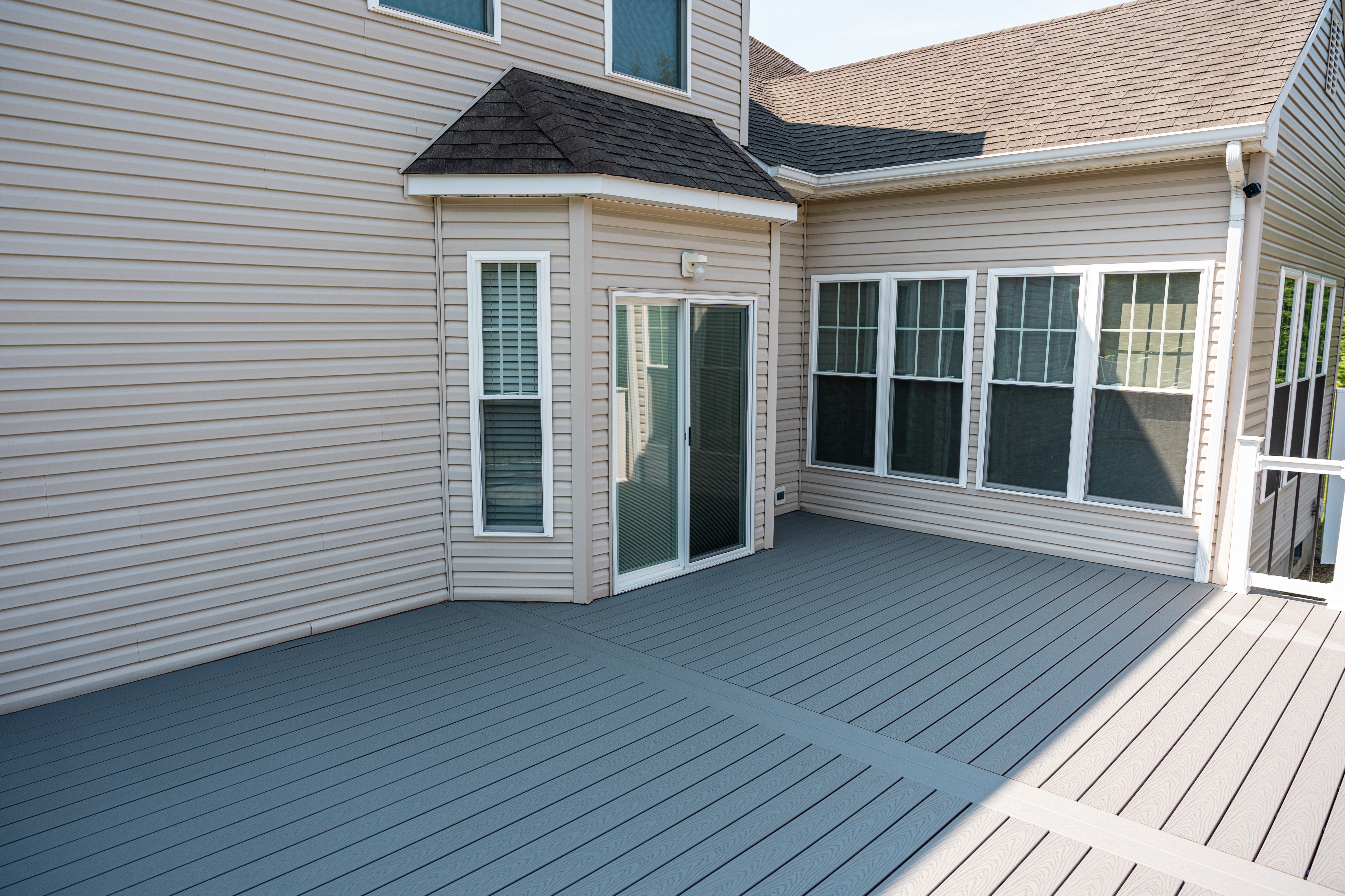 A spacious backyard patio with gray composite decking, bordered by beige siding of a house featuring multiple windows and a sliding glass door, under a clear blue sky.