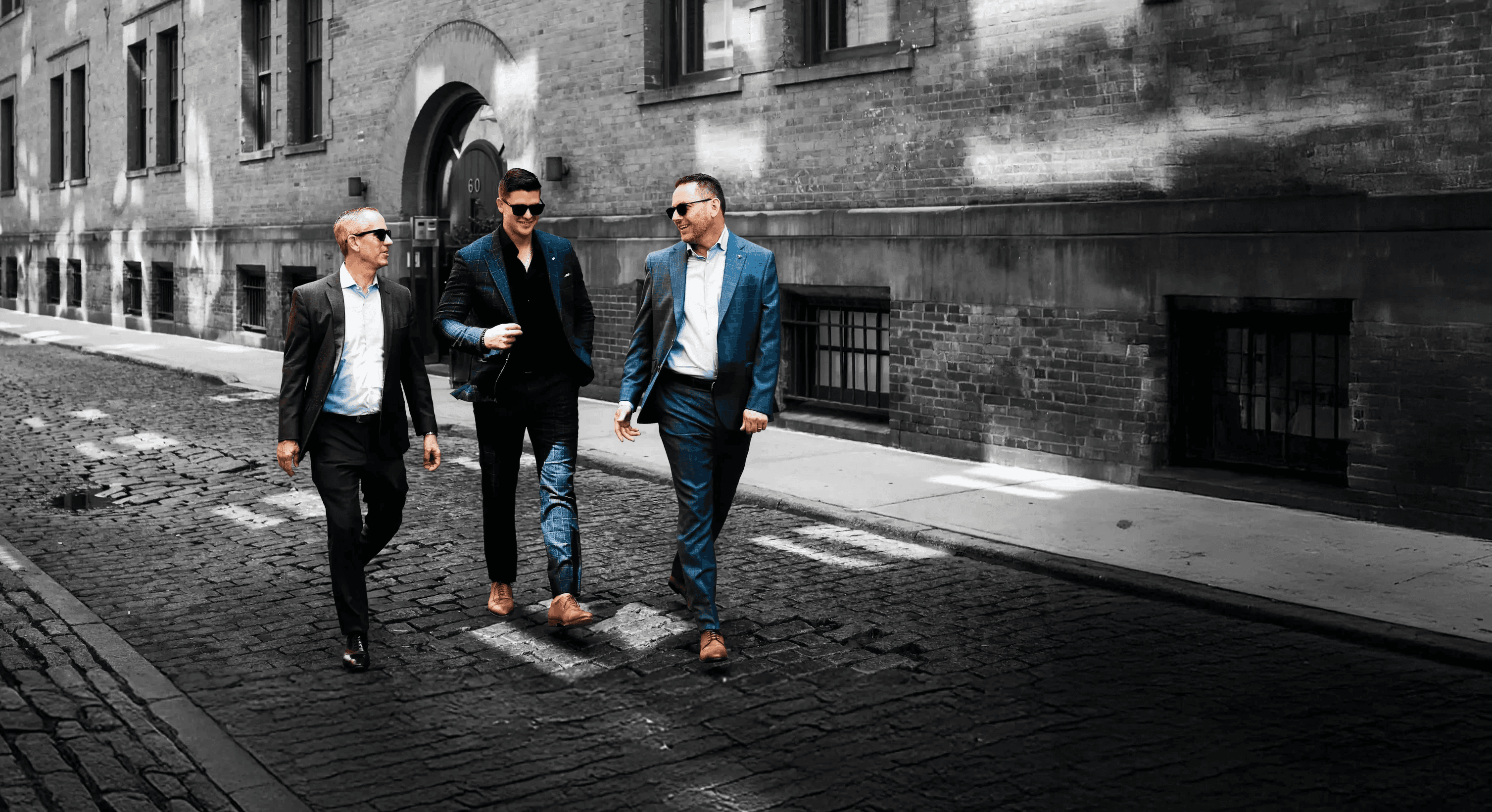 Three business professionals walk and talk along a cobblestone street beside a large building, dressed in smart casual attire.