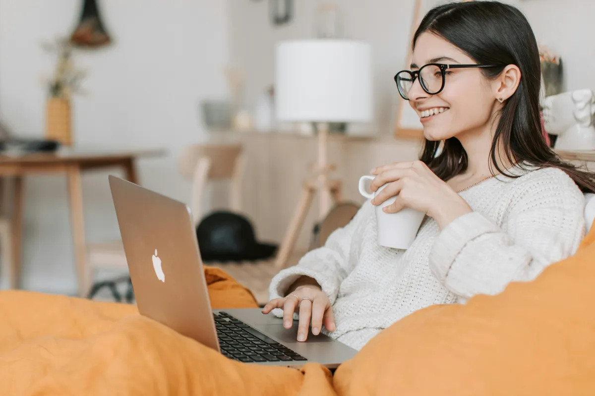 Young woman with glasses smiles while holding a mug and using a laptop on an orange couch.