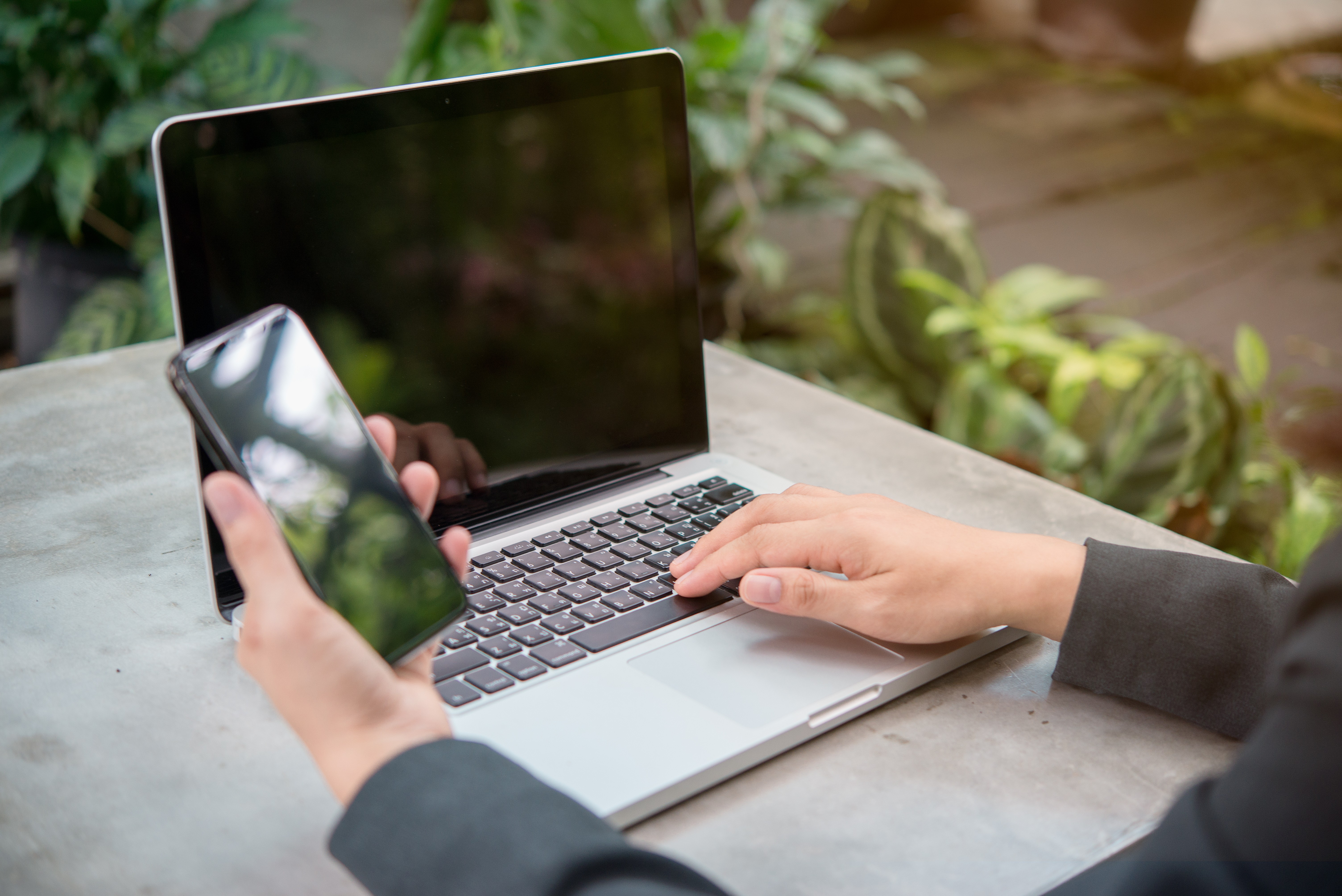 Hands using a phone beside a laptop in a leafy workspace
