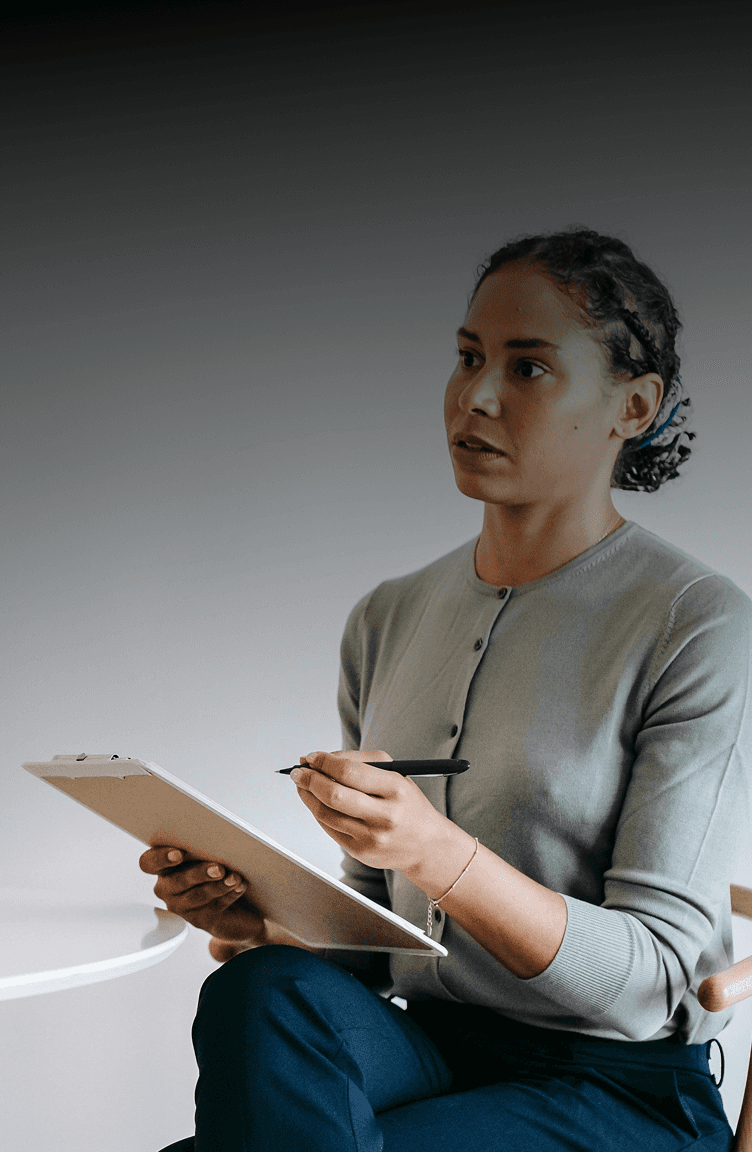 A woman with braided hair sits in a chair, holding a clipboard in one hand and a pen in the other. She is wearing a light gray cardigan and appears to be engaged in thought or discussion. The background is softly blurred, creating a focused atmosphere.