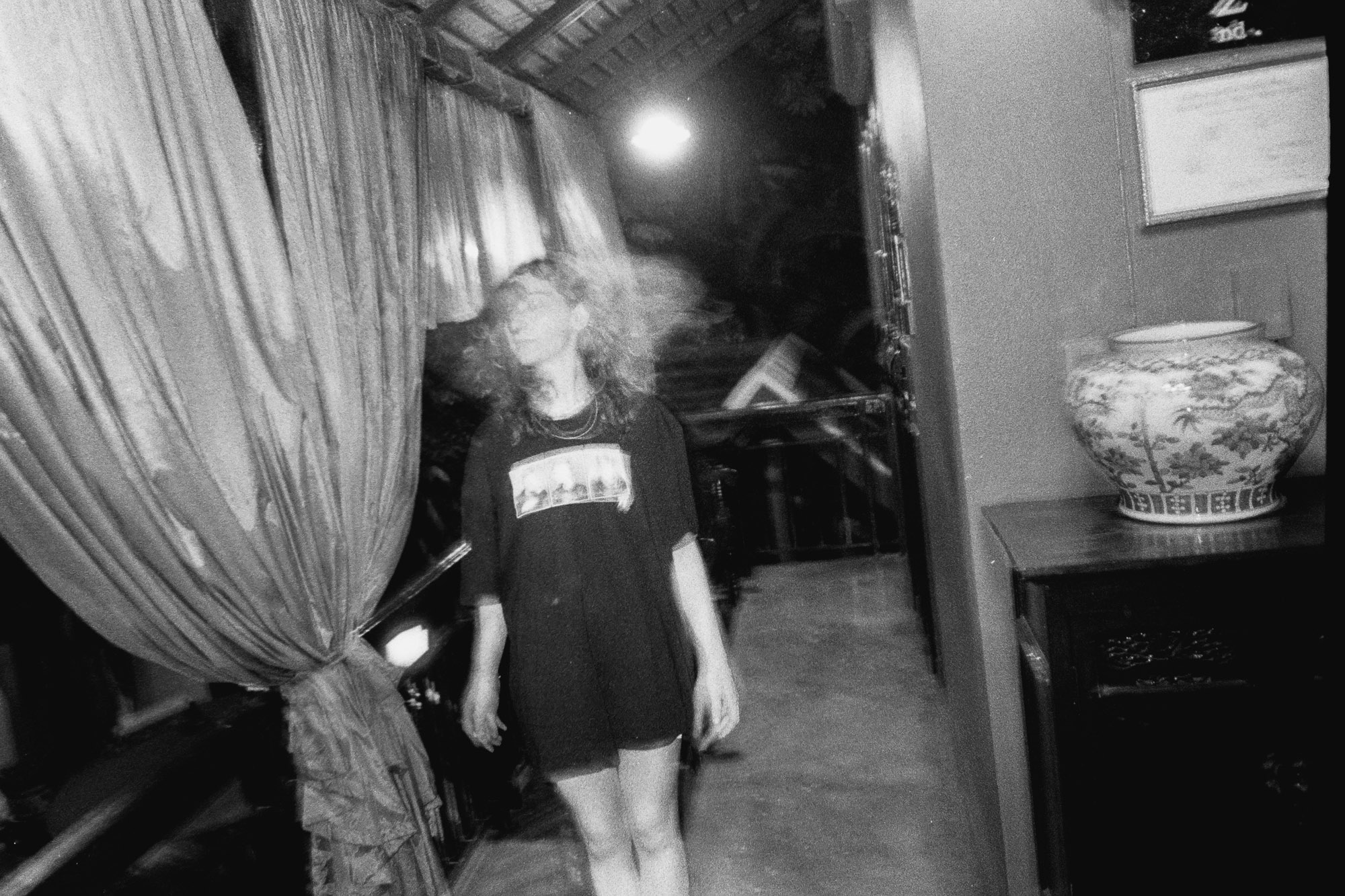 A person wearing a black t-shirt stands on a dimly lit balcony with sheer curtains blowing in the wind, next to a cabinet topped by a decorative vase.