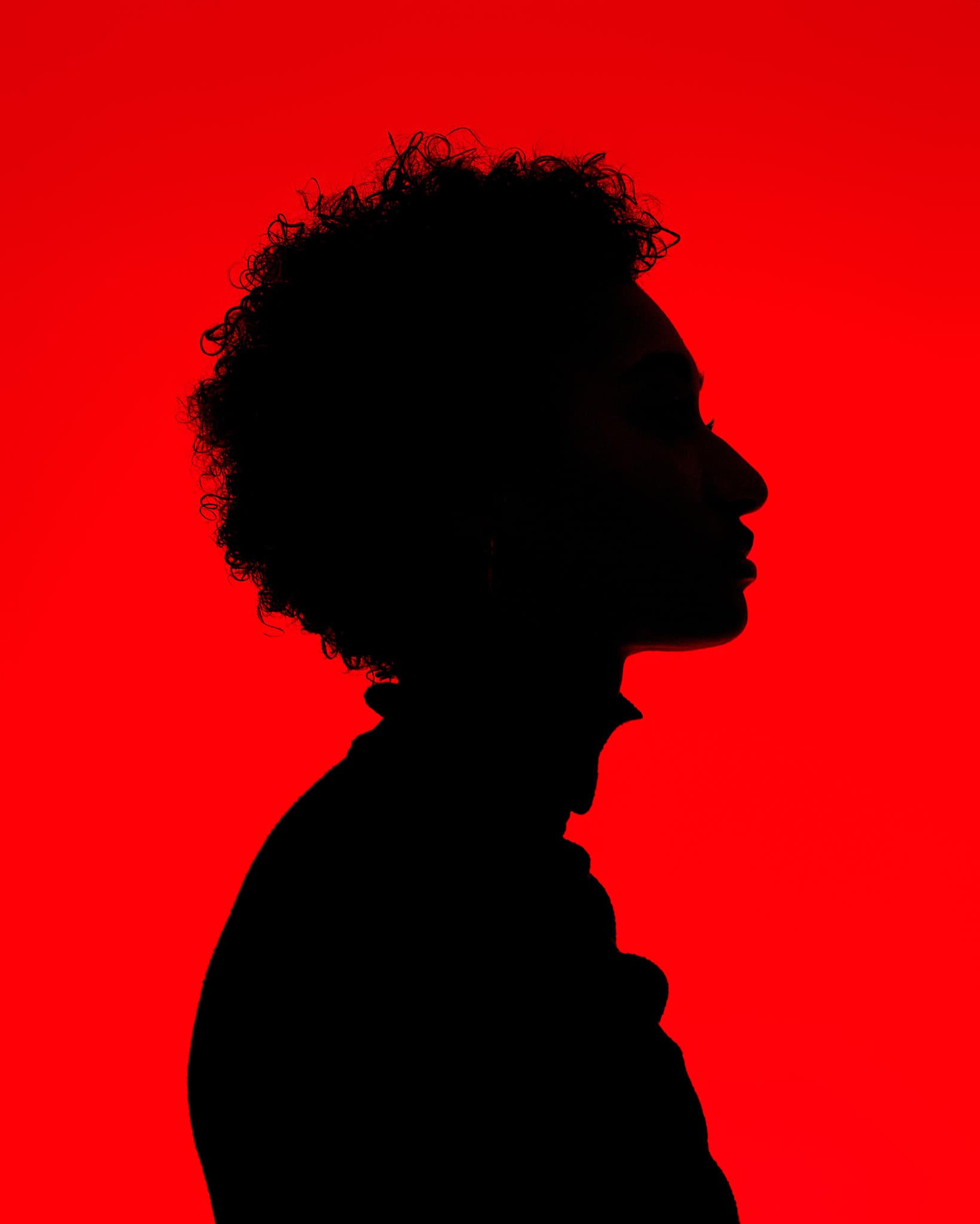 The profile silhouette of a model with curly hair against a red background in a natural light photography studio.