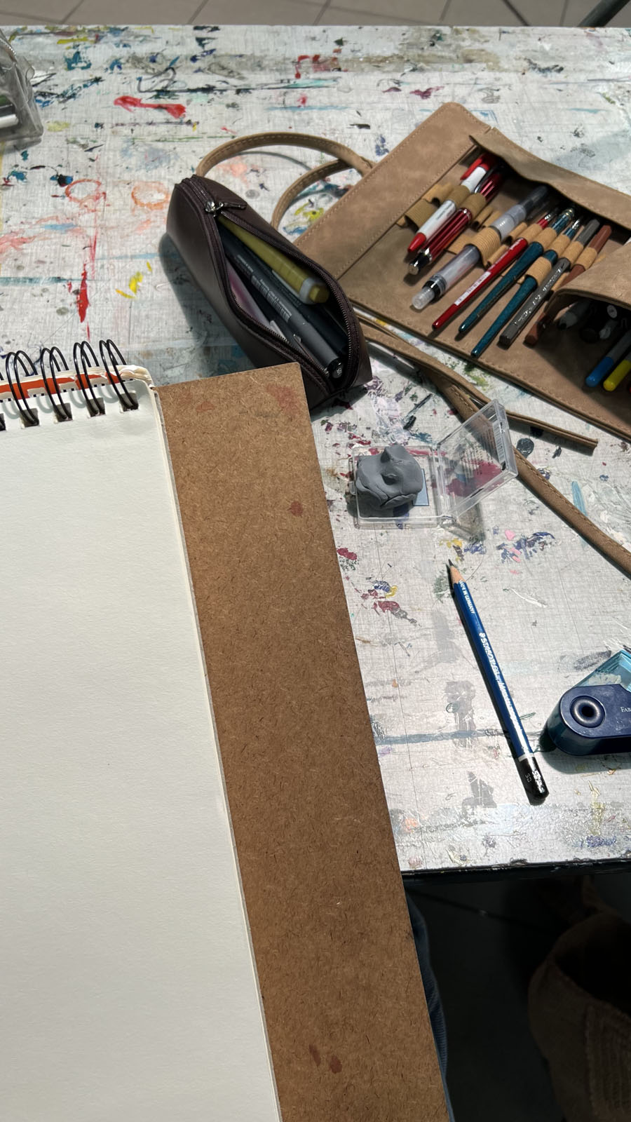 Een bovenaanzicht van een werktafel vol creatieve materialen. Er ligt een opengeslagen schetsboek met ringband, een etui met diverse pennen en stiften, een kneedgum in een plastic doosje en een blauw tekenpotlood. De tafel vertoont vele kleurrijke verfspatten en gebruikssporen.