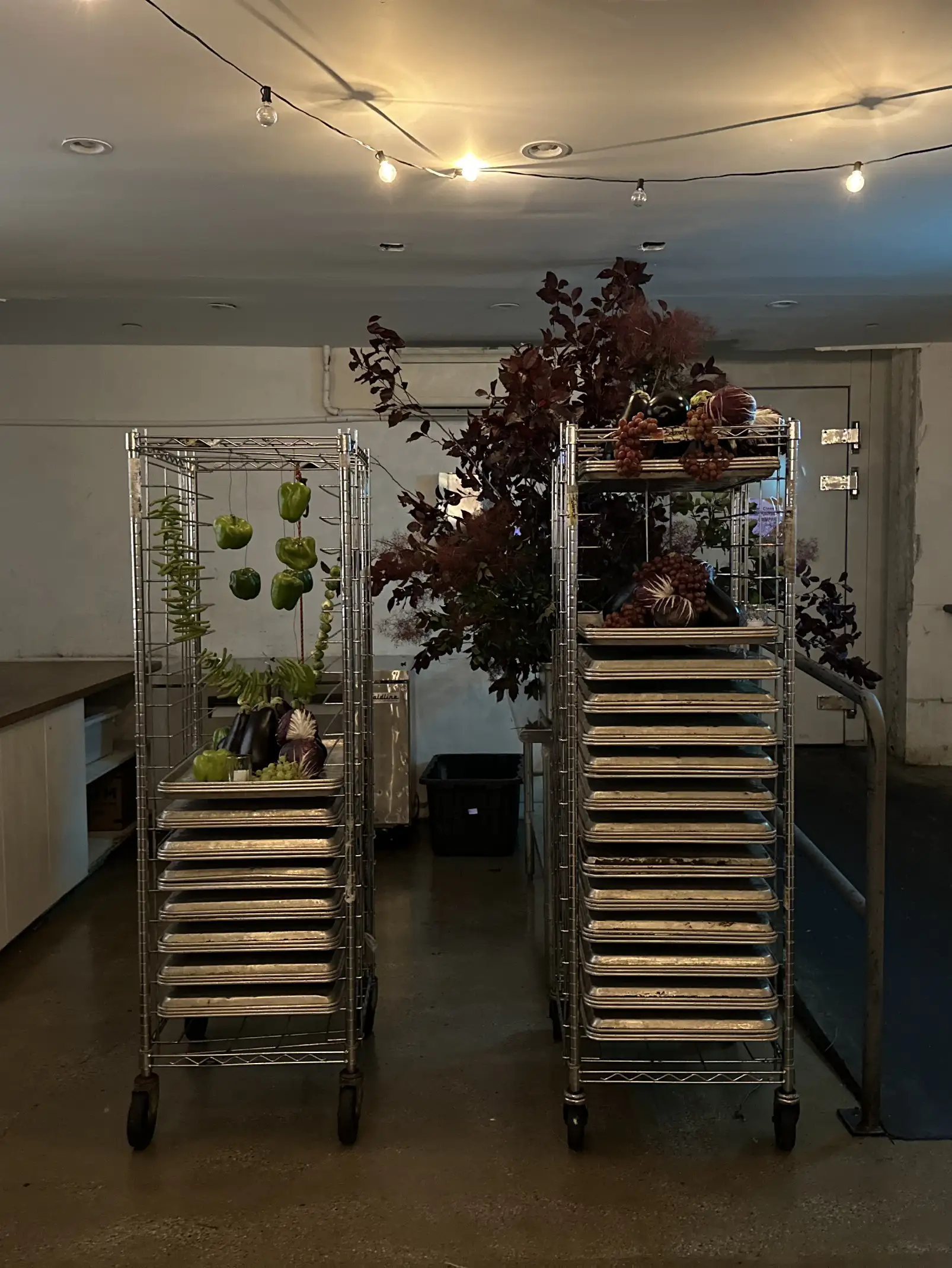 Dos estanterías metálicas con bandejas industriales decoradas con pimientos verdes, berenjenas, radicchio y ramas con follaje oscuro en una cocina iluminada con guirnaldas de luces.