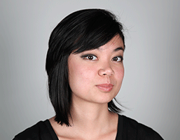A young woman with short, dark hair and bangs is captured in a head-and-shoulders portrait against a plain gray background. She wears a black top and looks directly at the camera with a neutral expression. Her skin tone is light, and her hair is styled in a straight, slightly angled cut.