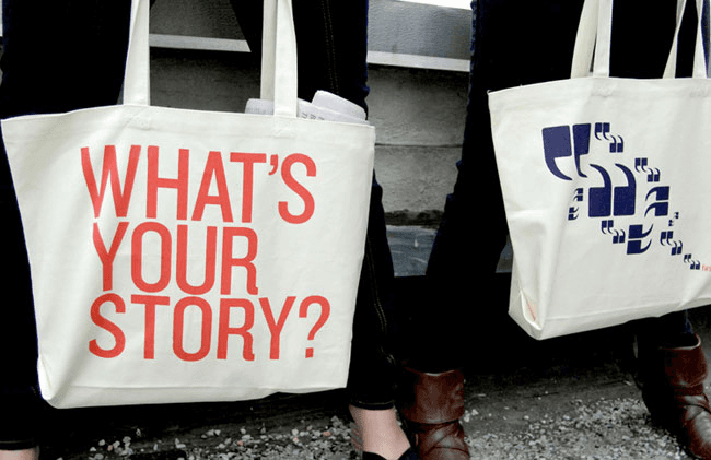 Two people are holding large white tote bags. The bag on the left has red text reading “WHAT’S YOUR STORY?” The bag on the right has a blue design of quotation marks. The bags are held in front of their lower bodies, obscuring their clothing. The background includes a gray wall and part of a staircase. The ground is covered with small stones or gravel.