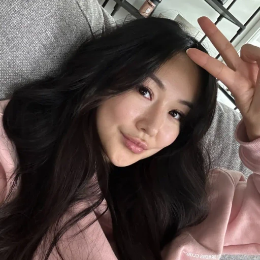 A person with long, dark hair is wearing a pink hoodie and making a peace sign with their fingers, while smiling at the camera in a cozy, indoor setting.