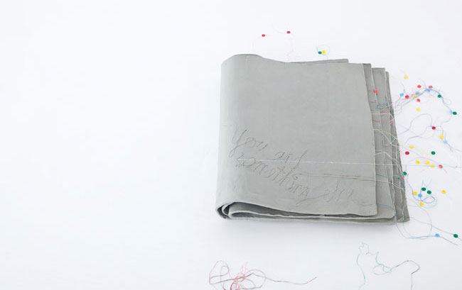 A gray fabric item, possibly a piece of clothing or blanket, lies on a white surface. The fabric has handwritten text in gray ink that reads “You and something” near the center. Multicolored pins are scattered around the fabric, some resting on the white surface and others near the fabric. The pins vary in color, including red, green, yellow, and blue. The white background provides a stark contrast to the gray fabric and the colorful pins.