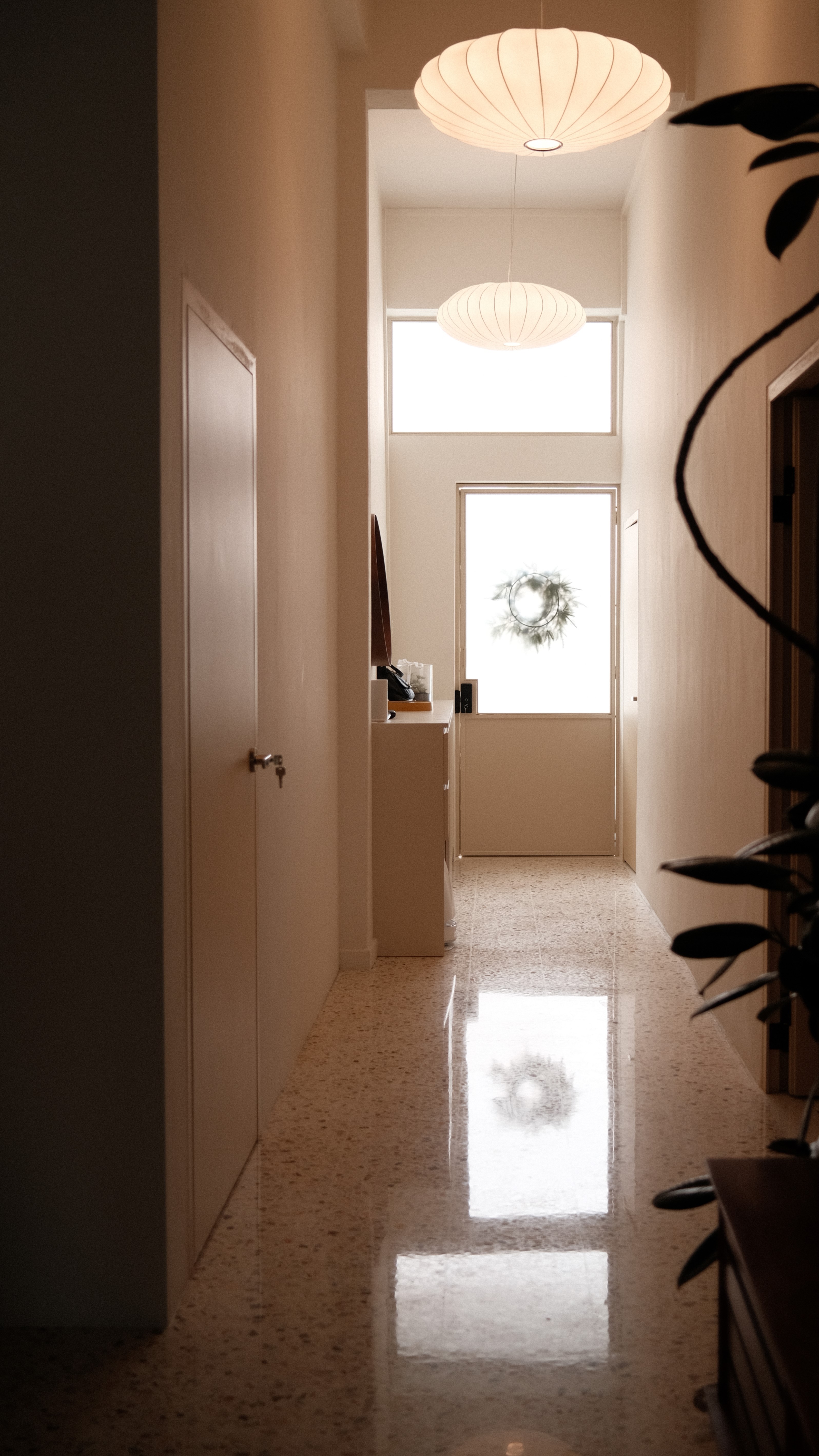 Bright hallway with terrazzo flooring, white doors, modern pendant lights, and a minimalist Christmas wreath on the glass door. Indoor plant in the foreground adds a touch of greenery.