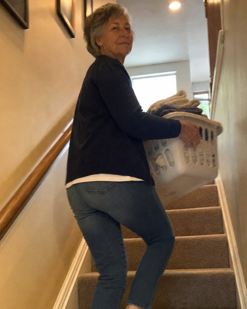A woman partway up her carpeted stairs carrying a full laundry basket and looking back over her shoulder, seen from the bottom of the stairs.
